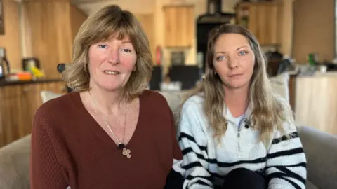 Sarah and Becky Armstrong sitting close to one another on a beige sofa in a caravan with a wooden kitchen behind them looking into the camera with neutral expressions.