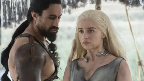 Warner Brothers Discovery Joe Naufahu and Emilia Clarke, characters from Game of Thrones, dressed in fantasy medieval leather clothing. They are scantily clad but there appears to be snow on the ground behind them. Their skin is dirty.