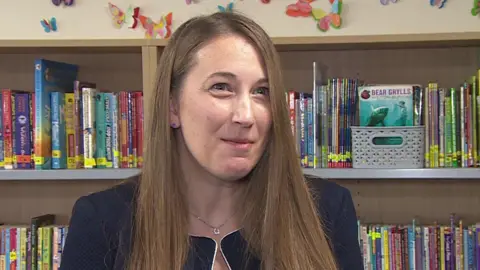 Sarah Simmons, the headteacher of Queen's Park Academy, Bournemouth, standing in the school library. A row of bookshelves is behind her.