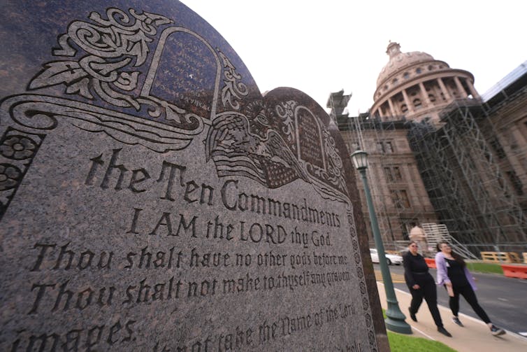 3 states are challenging precedent against posting the Ten Commandments in public schools Two women walk by an ornately carved stone monument, with a building with a large rotunda in the background.