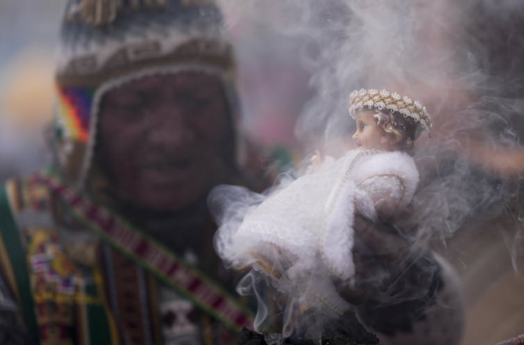 ‘Yes’ to God, but ‘no’ to church – what religious change looks like for many Latin Americans A man in a colorful knit hat and bright sweater or jacket holds up a small doll in a white robe that is surrounded by wisps of smoke.