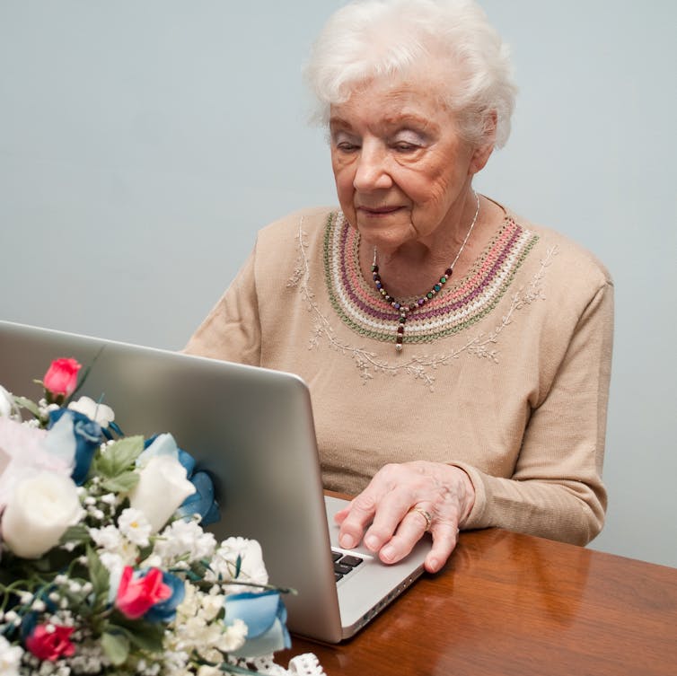 An elderly woman works on a laptop, looking grim, with a bouquet of flowers behind it.