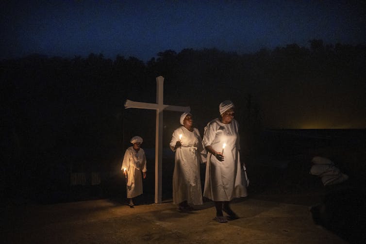 ‘Yes’ to God, but ‘no’ to church – what religious change looks like for many Latin Americans Three women in white robes and caps stand outdoors at nighttime by a large wooden cross.