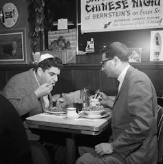 A black and white photograph of two men in suit jackets and Jewish head coverings seated at a table with signs on the wall.
