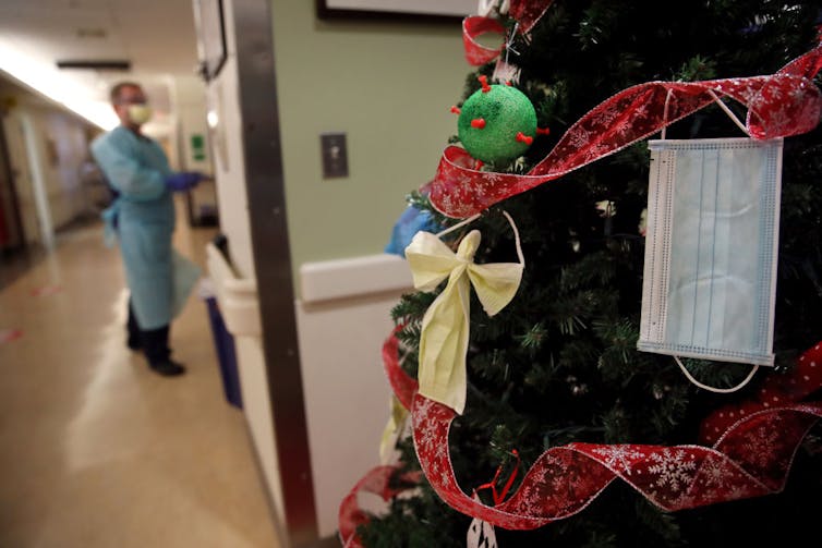 A man in blue protective equipment enters a room off of a corridor with a Christmas tree decorated in red ribbon, homemade ornaments, and a medical mask.