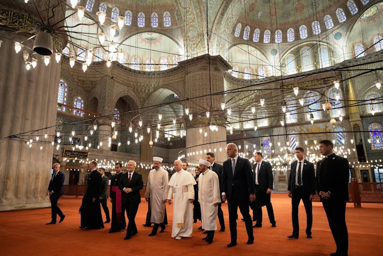 Pope Leo XIV’s visits to Turkey and Lebanon were about religious diplomacy Several people walking in a large dome-like structure with high ceilings. Some are dressed in black suits and a few in white robes.