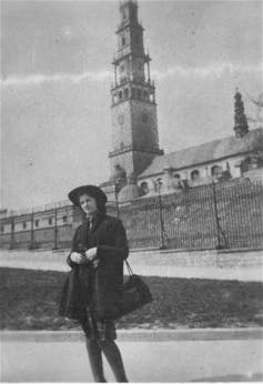 A black-and-white photo of a young woman in a coat and dark hat standing before a ramp leading up a building with a bell tower.