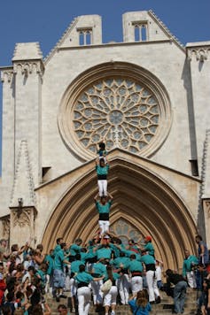 People in white pants and teal shirts form a human pyramid in front of a cathedral with a large, circular stained-glass window.