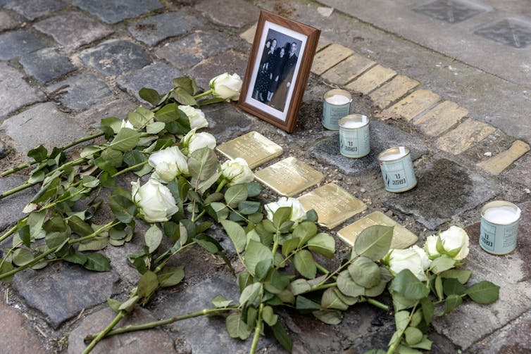 White roses placed on a sidewalk with four inlaid, bronze memorial stones, next to four candles and a framed family photo.