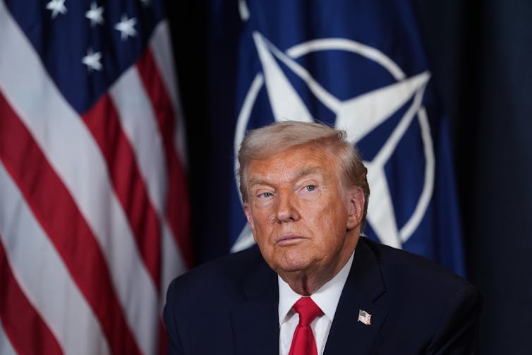President Donald Trump, wearing a blue suit and red tie, is seated in front of the American flag, with the NATO flag displayed beside it.