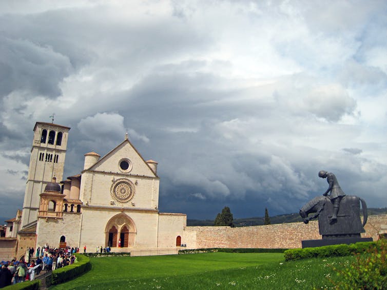 A Gothic-style cathedral sits under a cloudy sky as a line of people walks toward its entrance.