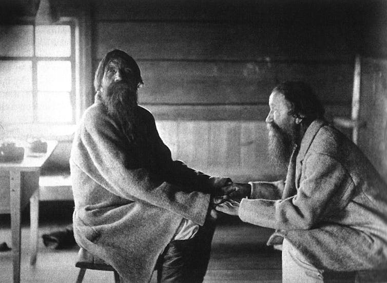 A black-and-white photograph of two men with beards sitting across from each other and holding hands.