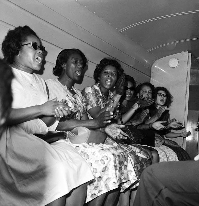 A black-and-white photo shows a row of seated women inside a van or small space clapping as they sing.