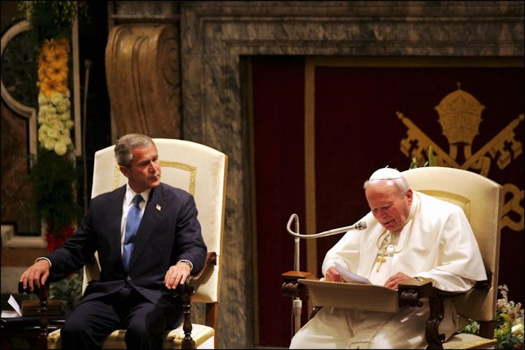 What is and isn’t new about US bishops’ criticism of Trump’s foreign policy A man in a suit and blue tie sits in an ornate white chair, next to another seated man in white robes reading into a microphone.