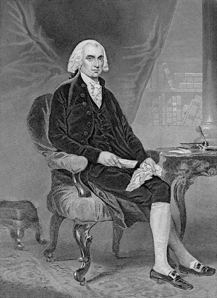 A stately man in a white wig, black waistcoat and knee breeches sits cross-legged beside a wooden side table.