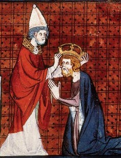 An illustration shows one man in a pointed hat putting a crown on a kneeling man's head, set against a red background.