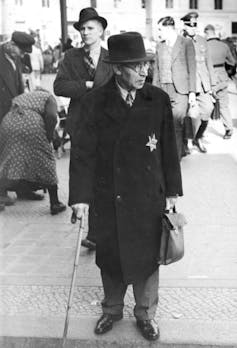 A man with a cane wears a top hat and black coat with a star badge on it.