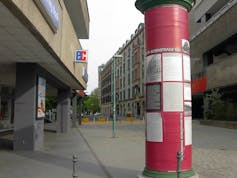 A large red pillar stands on a cobblestone street amid concrete buildings.
