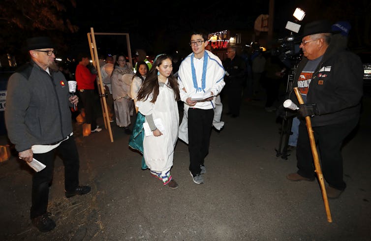 Young Latinos – and their commitment to social justice – are shaping the future of the Catholic Church A young man and woman, both of whom wear white costumes, walk at the front of a small procession outside at night.